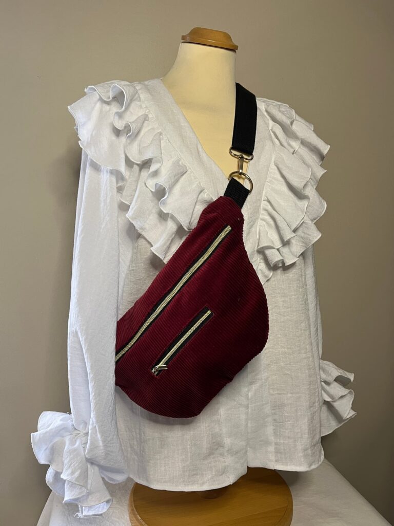 Sac banane grand format en velours côtelé bordeaux avec fermeture éclair dorée, laissant apparaître une doublure en tissu fleuri vintage.