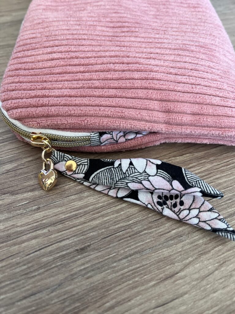 Gros plan sur la fermeture zippée, le breloque cœur et la tirette fleurie d'une pochette rose